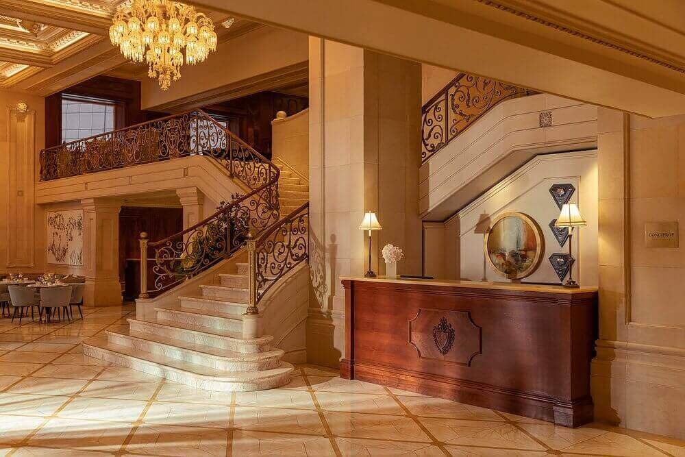 Elegant hotel lobby with chandelier, grand staircase, and ornate reception desk. Luxurious ambiance awaits.