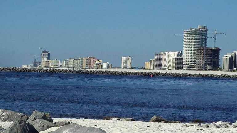 The image depicts a coastal landscape featuring a sandy beach with large rocks, a body of water, and a skyline of buildings including high-rise structures and construction cranes under a clear blue sky, keyphrase included.