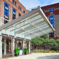 Modern building entrance with glass canopy, brick walls, columns, and lush greenery.
