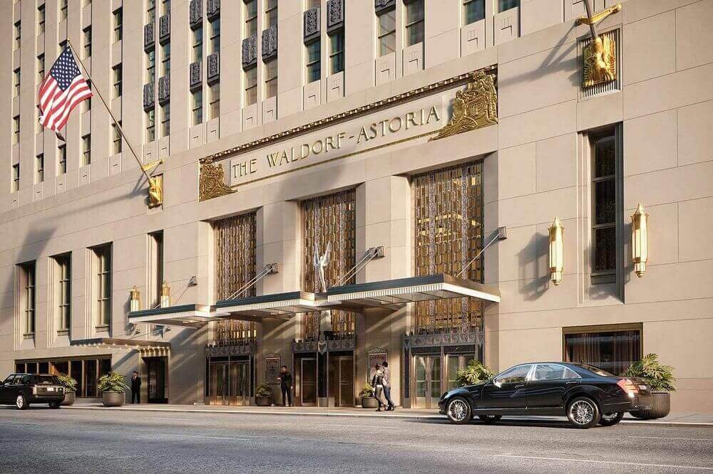 Elegant Waldorf-Astoria entrance with American flag, luxury cars, and ornate architecture.
